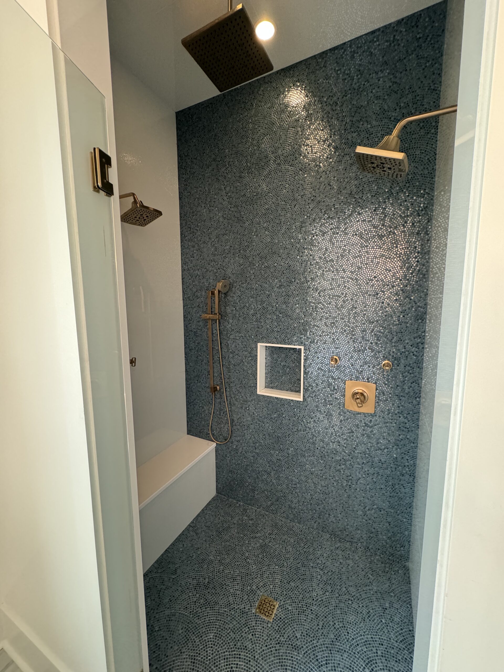 Custom tile shower remodel with blue mosaic tile wall, dual shower heads, glass enclosure, niche, and detailed finish work by Alabaster Contracting Solutions
